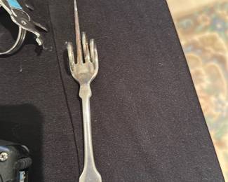Silverplate Pickle Fork — Heavy silverplate two-tine pickle/olive fork with a small turned handle and light surface wear consistent with age. Plain traditional pattern with no visible maker's marks in the photos; suitable for tabletop use or as part of a flatware lot. Shows minor scratching and patina but no bends or breaks. Measures small and lightweight, good for serving condiments.