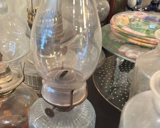 Glass Finger Lamp with Handle — Small clear pressed-glass finger lamp with molded petal/teardrop pattern and a single loop handle. The lamp has its original font, brass/silvered burner and a chimney; wick is present but appears used. Good decorative condition with expected surface wear and patina to the metal burner; glass has no obvious cracks visible in photos. Great as a display piece or for collectors of Victorian-style oil lamps — compact size shown in images.