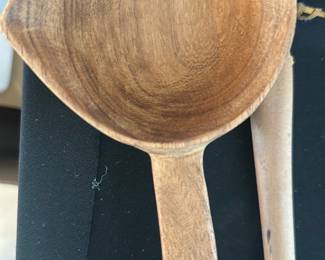 Hand carved wooden ladle — Hand-carved wooden ladle with a long tapered handle and shallow rounded bowl. Solid hardwood shows visible grain and wear consistent with age and use; finish is matte with a few dark spots and light surface abrasions. Simple utilitarian piece suitable for serving or display in a rustic or farmhouse kitchen; lightweight and well balanced by hand. Condition is good overall with expected surface wear but no visible cracks or breaks.