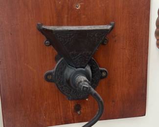 Wall-Mount Coffee Grinder — Decorative wall-mount coffee grinder with cast-iron body and wooden handle mounted on a small wood plank. The cast-iron hopper and ornate detailing show period styling and the curved crank ends in a turned wood grip; a shallow metal catch tray sits below. Overall compact and practical for a sun room display; handle cranks smoothly though grinder has surface oxidation and patina consistent with age. Good condition structurally with cosmetic wear, surface rust, and a few small screw/wood blemishes from mounting.