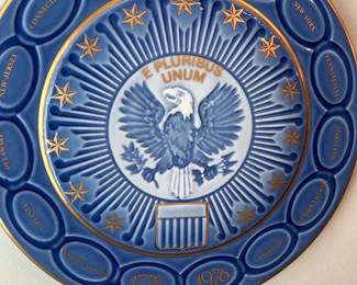 Bicentennial Eagle Decorative Plate — Decorative blue ceramic commemorative plate featuring a central eagle and "E PLURIBUS UNUM" motif, marked with the years 1776–1976 around the rim. The piece lists U.S. states in raised oval panels around the border and is trimmed in gold-painted accents. Approximately 12 inches diameter, suitable for wall display in the living room; lightweight and easy to hang with the existing plate hanger. Good condition overall with light surface wear to gold paint consistent with age and display.