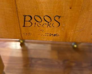 Boos Block Butcher Block Table — Boos Block butcher-block table with thick end-grain top and turned legs on brass caster cups. The maker stamp "Boos Block" is visible on the side; top shows expected cutting wear, stains and surface patina consistent with kitchen use. Solid construction typical of Boos cutting blocks makes it useful as a prep or island piece; top may benefit from sanding and refinishing. Overall sturdy but shows honest wear and cosmetic marks as photographed.