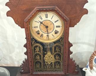 Wood Mantel Shelf Clock — Decorative wooden mantel clock with carved crest and octagonal bezel featuring a painted dial with Roman numerals and a gilt pendulum bob. The front glass has a black and gold painted scene; case shows age-related wear, surface scratches, and light discoloration on the dial. Likely late 19th–early 20th century style shelf clock with mechanical movement (movement not tested). Solid wood case with stepped base measures modestly for display and would suit restoration or decorative use.