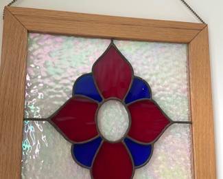 Stained Glass Floral Panel — Decorative stained glass panel featuring a red and blue floral motif set in textured clear glass with a light oak frame. The piece hangs by a metal chain and is ideal for a sun room window or wall display, adding colorful light when placed in direct sun. Overall framed dimensions are approximately 16 inches square, making it an easy-to-hang accent. In good condition with intact glass and frame, minor surface wear consistent with normal display use.