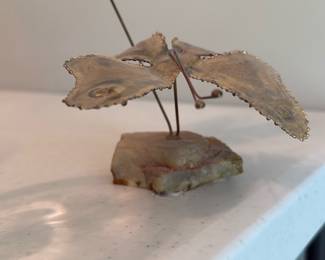 Brass Flower Sculpture on Stone Base — Decorative brass flower sculpture mounted on a rough stone base, shown in photos from multiple angles. The piece features textured, petal-shaped brass leaves with scalloped edges on thin brass stems; the stone base appears naturally formed with iron-staining and a flat underside for display. Good decorative condition with surface patina, light tarnish, and minor age wear consistent with vintage metal decor. Compact tabletop size makes it suitable for a shelf, mantel, or bedside table.