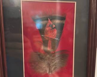 Cardinal Feather Print Framed — Framed artwork featuring a red cardinal perched on a feather against a deep red background. The piece is double-matted (cream inner mat, black outer mat) in a dark wood frame with glass. Good decorative piece for living room display; frame shows light surface wear and the glass has some reflections but no obvious cracks. Overall condition is good and ready to hang.