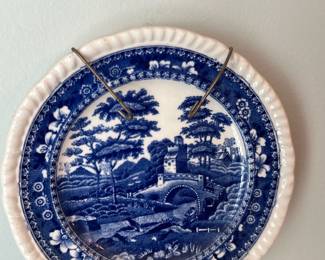 Blue Transferware Decorative Plate — Blue transferware decorative plate with classic pastoral bridge-and-cottage scene and scalloped rim. The plate is finished in blue on white earthenware with a molded fluted edge and is marked on the back with a blue oval impressed mark (see photos). Measures approximately 8 inches diameter and is suitable for living room wall display or cabinet decor. Good estate condition with minor surface wear and light crazing consistent with age; mounted on a metal plate hanger in photos. A charming example of blue transfer-printed earthenware popular for country-style décor.