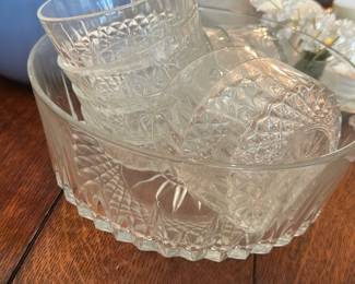Salad Bowl and 8 Bowls Clear Glass — Set of a clear glass salad bowl with eight matching clear glass serving bowls, perfect for family meals or entertaining. The pieces feature pressed-glass starburst and diamond-cut patterns with scalloped bases and sturdy rims, all shown photographed in the living room. Overall dimensions are modest and practical for tabletop use; see measurements for approximate sizing. In good, usable condition with light surface scratches from normal use and no visible chips or cracks in the photographed pieces.
