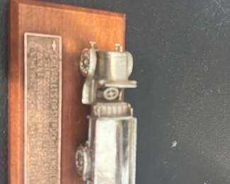 Cast Metal Pickup Truck Plaque — Small cast-metal pickup truck mounted on a wood plaque with a metal dedication plate. The piece appears decorative, likely a commemorative or recognition award; the truck measures only a few inches and shows light surface wear and tarnish consistent with age. Wood plaque has intact finish with minor scuffs. Good display piece for a shelf or desk; see photos for condition and plaque text detail.