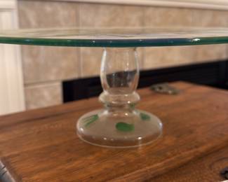 Hand-Painted Glass Cake Stand — Hand-painted glass cake stand with a round platter and short pedestal foot featuring colorful floral motifs in yellow, purple, pink and green. The painted decoration is visible on both the top surface and faint accents on the base; the clear glass is thin and delicate. Good estate condition with light surface wear and no visible cracks or chips in the photos. Useful as a serving piece or decorative centerpiece for a table.