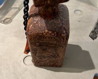 Tooled Liquor Decanter — Decorative tooled leather-covered perfume or scent bottle with a fitted stopper and attached chain; all sides show ornate floral carving and a figural cartouche on the front. The piece appears hand-tooled with warm brown leather over a rigid form and shows light surface wear and patina consistent with age; stopper fits but finish has scuffs. Compact accent sized decorative bottle suitable for display on a dresser or shelf; see photos for condition and details.
