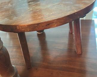 Child’s tray — Small oval wooden three-leg stand with a removable pegged-leg assembly; looks handmade or early 20th-century country style. The top shows surface wear, scratches and finish loss consistent with age; legs are pegged into recessed slots on the underside and one peg area shows wear but appears intact. Solid, lightweight construction suitable for displaying plants, a small lamp or serving as a riser; measures small and easy to move. Good usable condition with visible cosmetic wear and patina; no loose major breaks seen in photos but some edge and surface scuffs.