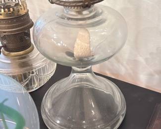 Clear Glass Oil Lamp — Tall clear glass oil lamp with a round reservoir, replacement cotton wick, and fitted glass chimney. The metal burner shows age-appropriate patina and secures the chimney with three hinged clips; chimney is intact with no visible cracks. Good decorative or functional piece after cleaning and filling; measures roughly 16 inches tall (see dimensions).