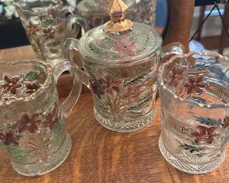 Pressed Glass Floral Creamer+Sugar Set — Three-piece pressed glass cream and sugar set with lid, featuring molded floral medallion decoration hand-painted in pink and green with gilt highlights on the lid finial. The set includes a small creamer, a lidded sugar jar, and an open tumbler/compote each with scalloped rims and textured bases; the painted decoration shows light wear to gilt and paint from use. Good usable condition with normal surface wear and a couple small nicks possible on rims (see photos); a charming decorative set for dining-room display or everyday use.