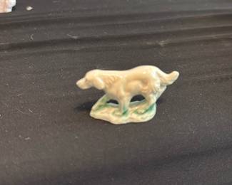 Miniature Dog Figurine — Small glazed ceramic dog figurine depicting a hunting or retriever-type dog standing on a grassy base. Neutral cream glaze with green-tinted base; hand-painted details are minimal. Appears to be a small shelf or curio piece with light surface wear and no visible chips or cracks in provided photos. Ideal as a small decorative accent for a curio shelf or collection of porcelain animals.