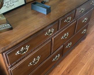 Wooden Nine-Drawer Dresser — Classic wooden nine-drawer dresser with attached mirror, offering generous bedroom storage and traditional brass-tone hardware. Constructed of stained wood with molded drawer fronts and dovetailed-style drawer boxes (appears plywood interior) in a medium brown finish. Measures approximately 60 inches wide, 34 inches tall (including mirror base height to top of dresser surface 34 in), and 18 inches deep; top shows light surface wear and drawer interiors are clean. Overall in good condition with surface scratches and light finish wear consistent with normal use; hardware intact and drawers functional.