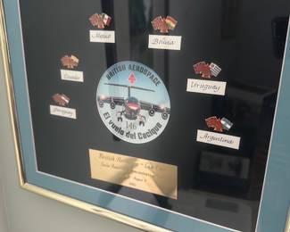 British Aerospace Framed Plaque — Framed British Aerospace commemorative plaque from the 1990 Latin American demonstration tour, titled "El vuelo del Cacique" and featuring a central aircraft decal and enamel country pins labeled Mexico, Bolivia, Ecuador, Paraguay, Uruguay, and Argentina. The piece is double-matted in teal and black with a gold metal nameplate and housed in a gold-tone metal frame — photographed in the office room. Overall good condition with minor wear to the frame and glass surface from display; matting and pins appear intact and original. A distinctive aviation display item for a collector or office wall, ideal for aviation enthusiasts or corporate memorabilia collections.
