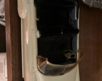 Honeydew Convertible Car Bank — Metal toy convertible coin bank in cream with black interior and clear plastic windshield. The underside shows a coin slot and winding/key mechanism; some surface wear and minor paint loss at edges are visible. Good collectible condition for display or shelf use as a vintage-style metal car bank; measures small and light enough for tabletop placement. No maker name legible in the photos.