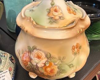 R & S Covered Floral Jar — Decorative covered porcelain jar marked Royal Rudolstadt on the base, featuring hand-painted yellow-orange roses with soft green shading and gilt accents on a cream ground. The piece stands on four small molded feet and has a scalloped lid with a finial, showing delicate brushwork and applied gold trim typical of late 19th–early 20th century German porcelain. Overall condition is good with light wear to gilt on edges and no visible chips or cracks in photographed views; base shows the factory mark. Measures modestly and is ideal as a decorative accent or for collector display.
