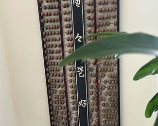 Large Wooden Chinese Abacus Wall Art — Large decorative Chinese abacus displayed as wall art with carved ivory-colored characters on a central black panel. Constructed from wood with multiple vertical rods of wooden beads and a slim rectangular frame, this piece hangs in the sun room and serves as both a folk-art display and conversation piece. Overall size is estimated to fit a tall narrow wall; see dimensions for specifics. Good vintage condition with natural wear to beads and finish consistent with age, no major damage visible.
