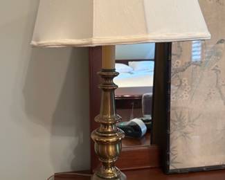 Brass Table Lamp with Shade — Stately brass table lamp with classic turned column and stepped round base, shown with a tapered fabric shade. The lamp appears to be a solid heavy brass construction with a faux candle sleeve at the socket and a standard turn-knob switch; shade shows light wear at edges. Works with standard bulbs (shown with LED bulb) and is well suited for bedroom or living-room use — small surface scratches and patina consistent with normal use are visible on the base.