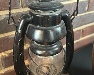 Dietz Embury Railroad Lantern — Classic black railroad-style lantern marked "Embury MFG CO" on the base with glass globe and metal burner insert. Solid metal construction with bail handle and protective wire around the globe; shows surface wear, scuffs, and light paint loss consistent with age but structurally sound. Decorative or functional as a display piece; glass appears intact with no visible cracks. Measures compactly and is a recognizable example of late 19th/early 20th-century style railroad lanterns.