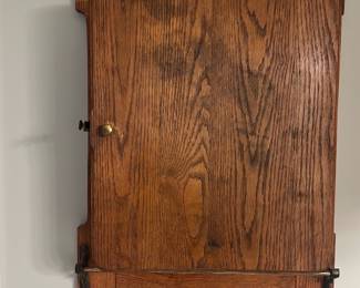 Antique Wall-Mounted Oak Medicine Cabine — Solid oak wall-mounted medicine cabinet with two internal shelves and an exterior hinged door; pictured in the master bedroom. Finished in a warm medium oak stain with brass knob and metal support hardware, the cabinet offers shallow storage for bathroom or bedroom essentials. Measures approximately 24 inches tall x 16 inches wide x 6 inches deep and mounts directly to the wall for easy installation. In good vintage condition with light surface wear and patina consistent with age; hinges and latch functional but show some oxidation on metal parts.