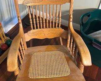 Oak Spindle Back Rocking Chair — Solid oak spindle-back rocking chair with carved crest rail and turned stretcher details; the seat features a woven cane seat insert. Sturdy construction and classic country styling make it a versatile piece for a living room or nursery. Shows expected surface wear, light scratches and finish dulling consistent with age, and the cane insert appears intact but may be a newer replacement. Measures under standard chair size; see dimensions and notes for specifics.