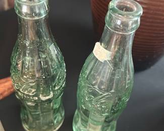 Coca-Cola Embossed Mini Bottle — Two small embossed Coca-Cola glass bottles in light aqua-green glass; both show the classic contoured bottle shape with embossed Coca-Cola script and light surface wear consistent with age. One bottle retains an old paper price sticker; no visible chips or large cracks in the photos but bottles show scuffs and surface residue and would benefit from a gentle clean. Good decorative pair for collectors of soda memorabilia or vintage glassware; compact size shown in photos.