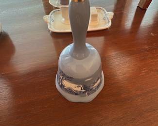 Blue Porcelain Dinner Bell — Small blue porcelain hand bell with a painted winter cottage scene around the skirt and a gilt tip on the handle. The bell is solidly made ceramic with a glossy glaze and shows light surface wear consistent with display; no visible chips or cracks in photos. Attractive as a decorative piece or table accent for a dining room collection; measures small enough for shelf display or to ring by hand.