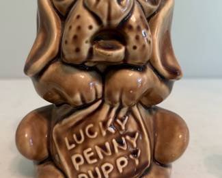 Lucky Penny Puppy Ceramic Bank — Small brown glazed ceramic puppy bank marked "LUCKY PENNY PUPPY" on the front. Cute figural coin bank in a mottled brown glaze with black-painted eyes and simple molded details; appears to be a mid-century style novelty bank. No visible brand or maker marks in the photos; condition is good with normal surface wear and light glaze crazing visible in close-up images. Compact size suitable for shelf display or children's room decor.