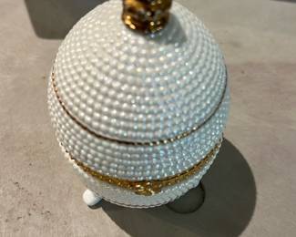 White Beaded Trinket Box — Small decorative white beaded trinket box with a hinged lid and gold-tone metal trim. The exterior is covered in rows of tiny pearlescent beads and it sits on three small feet; the interior shows a smooth pale lining and an ornate gilt rim around the opening. Good decorative condition with normal light handling wear to gilt trim visible in photos; hinge appears intact and the lid closes properly. Compact accent piece suitable for jewelry or small keepsakes.