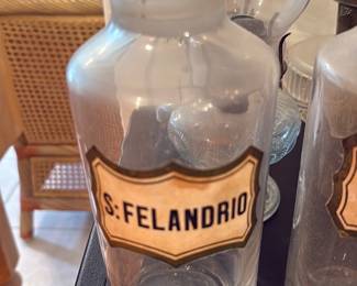 Glass Apothecary Jar — Tall clear glass apothecary jar labeled "S: FELANDRIO" on a paper or applied label; the jar includes a fitted glass lid with a raised knob and shows typical surface wear and light residue from use. The cylindrical form and label suggest a vintage pharmacy or apothecary storage piece suitable for display or decor; the glass is intact with no visible cracks or chips in the photos. Stands approximately the height shown relative to nearby objects; good vintage condition with shelf wear and label age.