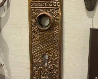 Ornate Brass Door Backplate — Decorative cast brass door backplate with intricate floral and geometric relief detailing and two keyhole openings plus a round knob bore. Solid-feeling hardware piece likely from an early 20th-century or reproduction set; the warm brass finish shows surface wear and patina consistent with age and use, including small nicks and finish loss around the knob hole. Good candidate for restoration on a period interior or for collectors of architectural salvage. Overall slim rectangular profile; see dimensions for fit.