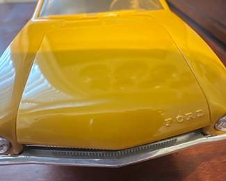 Yellow Ford Car Model — Bright yellow diecast Ford model car displayed in an office room; compact collectible suitable for display. Solid metal body with plastic windows and interior, chrome-tone bumpers and hubcap detailing, appears to be a 1:18–1:24 scale promotional or toy replica. Overall length is small enough for shelf or desk display and it's in good condition with light surface wear and minor scuffs consistent with handling. No visible broken parts; wheels and trim intact though finish shows small imperfections from age or play.