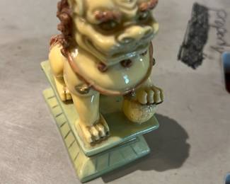 Ceramic Foo Dog Figurine — Glazed ceramic foo dog / Chinese guardian lion figurine with a pale yellow-green glaze and brown-detailed mane. The piece sits on a stepped rectangular base and the front paw rests on a textured ball; glaze shows a glossy finish with light surface wear visible in photos. A decorative accent for a shelf or mantel, solid but lightweight ceramic construction; no obvious large chips or repairs seen though close inspection recommended for hairline crazing typical of glazed pottery.