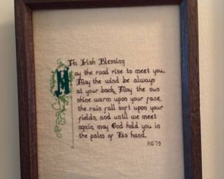 Irish Blessing Framed Cross-Stitch — Hand-stitched framed piece featuring the poem titled "The Irish Blessing" worked in green and brown threads on a neutral evenweave ground. The sampler includes a decorative illuminated initial and is signed/dated "RG '79" in the lower right; mounted in a simple dark wood frame with glass. Good overall condition with light age-appropriate toning to the fabric visible in the photos and no obvious tears or stains to the stitching or frame.