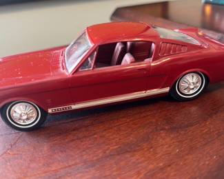 Red 1965 Ford Mustang Model Car — Collector-quality red 1965 Ford Mustang die-cast model presented as a detailed scale replica. Molded metal body with painted red finish, clear plastic windows, interior bucket seats and stamped wheel details; side "Mustang" script visible. Compact display piece roughly the size of a standard 1:24–1:32 model suitable for office or shelf display; minor surface dust and light paint wear visible. Overall good condition for a used collectible with no broken pieces noted and intact wheels and trim.
