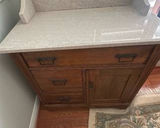 Oak Washstand with Marble Top — Solid oak washstand featuring a light gray marble top and backsplash, offering classic storage and a decorative presence in the living room. The piece has three left drawers and a right cabinet with original ornate brass drop handles and carved trim, finished in warm medium oak. Overall size is compact and suitable for hall or entry use; the marble top shows light surface marks from use. Good structural condition with normal surface wear and minor scratches consistent with age.