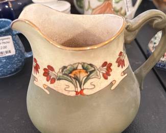 Olivetian Haynes Pitcher — Olivetian Haynes small ceramic pitcher with matte green glaze and hand-painted floral band under a gilded rim. Marked on the base “Olivetian HAYNES” (see photos), this decorative cream/jug displays light crazing and age-appropriate surface wear but no visible chips or cracks. Ideal for display or as a small serving pitcher, the molded handle and art-nouveau inspired painted motif give it decorative appeal. Seller notes list provenance as Olivetian Haynes and item was photographed in a living room setting.