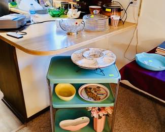 **Photo — Vintage Green Metal Rolling Cart & Retro Kitchenware** **Tags:** vintage 3-tier rolling cart, mid-century metal utility cart, California souvenir plate, marble relish dish, mid-century mod bowls, cake dome, footed glass punch bowl A retro three-tier green metal rolling cart is a fun mid-century find, topped with a marble-look divided serving dish. Its shelves hold a mustard-yellow mixing bowl, a vintage California souvenir plate, and colorful mid-century bowls with a vase of faux flowers. On the counter above, more kitchen collectibles round out the scene — a glass cake dome, footed punch bowl, and vintage teakettle.