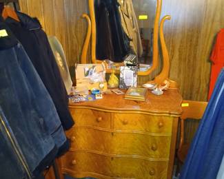 Photo — Antique Burl Wood Dresser with Swivel Mirror & Vintage Coats Tags: antique burl wood dresser, swivel mirror vanity, serpentine front chest, wooden picture frames, vintage men’s leather jacket, vintage coats, jewelry box A striking antique burl-wood dresser with a serpentine front, four dovetailed drawers, and an attached swivel mirror on curved supports — a real showpiece for a bedroom. Small collectibles are displayed on top, including picture frames, a jewelry box, and a shell dish. Nearby racks offer a selection of vintage coats and jackets, including a leather jacket and a lined denim coat.