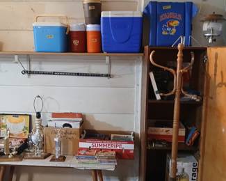**Photo — Coolers, KU Fan Gear, Elvis Calendars & Vintage Coat Rack** **Tags:** vintage coolers, Coleman cooler, water jugs, Kansas Jayhawks blanket, KU memorabilia, Elvis Presley calendars, wood coat rack, gas lantern, trophies, vintage records Camping and tailgate essentials up top: a Coleman cooler, vintage water jugs, ice chests, and a Kansas Jayhawks blanket — perfect for KU fans. Below, a fun mix of collectibles including vintage trophies, a box of Elvis Presley calendars ($1 each), records, and cookbooks. A wooden coat rack and vintage gas lantern round out the eclectic finds in this corner.