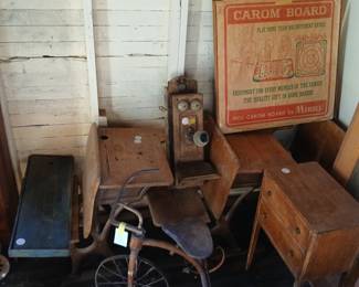 **Photo — School Desks, Antique Wall Phone, Tricycle & Vintage Game** **Tags:** antique school desk, wood ink well desk, oak wall telephone, crank phone, vintage tricycle, small dresser, Merdel Carom Board, vintage game box A wonderful lot for antique and collectible lovers: a wood school desk with ink-well cutout, an antique oak wall-mount crank telephone, and a rusty vintage tricycle. Behind them, a small wood dresser and a fantastic vintage #100 Carom Board by Merdel in original box add to the mix of primitive schoolhouse and childhood collectibles.