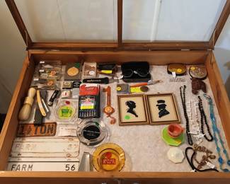 This display case is packed with small treasures — great for the “surprises in the basement” line. Photo — Display Case: Vintage License Plates, Silhouette Portraits, Jewelry & Advertising Ashtrays Tags: vintage Kansas farm license plates, straight razor shaving set, silhouette portrait art, vintage costume jewelry, Ramada Inn ashtray, Holiday Inn ashtray, 1941 Kansas chauffeur license, pocket knives, vintage eyeglasses A treasure trove of small collectibles: vintage Kansas farm license plates (1956–57), an antique straight razor and shaving brush set, and a pair of gilt-framed silhouette portraits. Advertising collectors will love the vintage motel ashtrays (Ramada Inn, Holiday Inn) and a rare 1941 Kansas chauffeur’s license/badge. Rounding it out — an assortment of vintage costume jewelry necklaces and bracelets, pocket knives, vintage eyeglasses, and small pocket lighters, all displayed in a glass-top wood case.