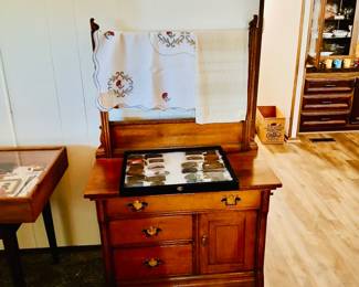 Antique Oak Washstand with Towel Bar & Belt Buckle Collection Tags: antique oak washstand, towel bar dresser, vintage embroidered linens, cross stitch towel, belt buckle collection display case, primitive chest of drawers A beautiful antique oak washstand with towel bar back, dovetailed drawers, and a cabinet door — a versatile piece for any bedroom or bath. Draped on top, vintage hand-embroidered cross-stitch linens add cottage charm. A collector’s display case of vintage belt buckles sits on the surface, appealing to Western and Americana collectors alike.