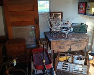 **Photo — Antique Door, Small Chest, Wire Buggy & Vintage Wagon** **Tags:** antique paneled door, small wood chest of drawers, wire doll buggy, zinc top table, vintage red wagon, tricycle, doll crib, Skelly oil cans A great architectural salvage piece — a solid wood paneled door — sits alongside a small chest of drawers and a rustic zinc-topped table. Nearby, the white wire doll buggy makes another appearance, along with a red vintage wagon, tricycle, and wood doll crib. Skelly Oil advertising tins are visible on the shelf above, tying together this room of primitive and childhood collectibles.