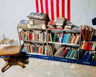 Home Library Collection, Antique Swivel Chair & American Flag Tags: vintage hardcover book collection, matched book set, home library shelves, vintage magazines, antique wood office chair, Pepsi crates, American flag  A substantial personal library fills these shelves — rows of hardcover fiction and reference books in varied bindings, with a matching set of maroon/burgundy volumes visible on the lower right shelf that looks like a complete reference or classics set. Stacks of vintage magazines and paperbacks top the shelves as well. A lovely find for book collectors, built over years by a former librarian and lifelong reader. An antique wood swivel office chair with claw feet sits ready alongside for browsing in comfort.
