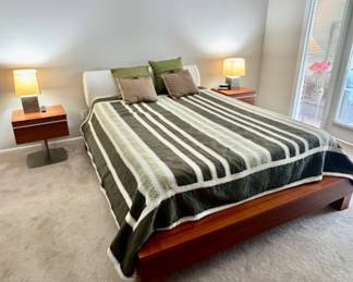 Mid-century modern style bedroom suite, featuring a low-profile wood platform bed with contrasting white headboard. Flanked by the same two floating wooden pedestal nightstands and square ambient table lamps.