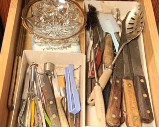 52 - Drawer Lot of Assorted Items (Description): Drawer lot containing an assortment of kitchen items including various utensils such as whisks, spatulas, knives with wooden handles, a slotted spoon, peelers, and other cooking tools. Also included is a pressed glass dish with scalloped gold-trimmed rim and a ceramic spoon rest with decorative text. Measurements not recorded.